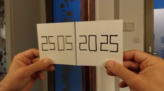 A hand holding a paper on which 2025 is written perpendicular to a mirror. With the symmetry, it reads 25 05 2025.